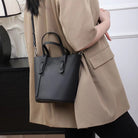 Person wearing a beige coat holding a black handbag against a neutral background