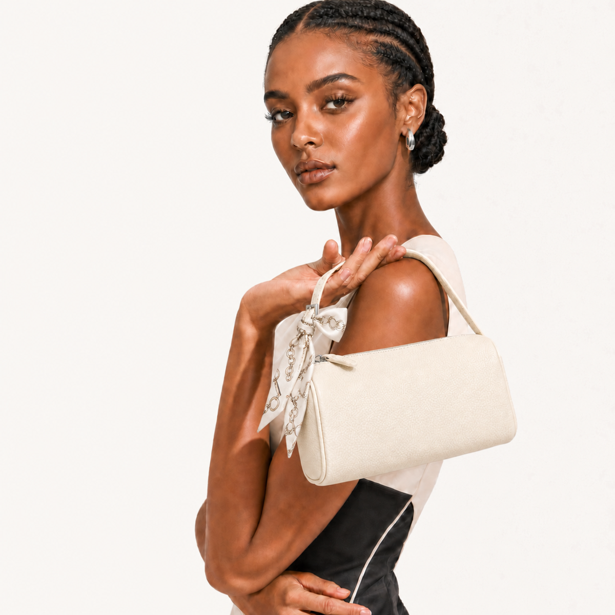 A model with braided hair holds a cream-colored structured shoulder bag with a matching fabric tie detail. She is wearing a black and white top against a clean, off-white studio background.