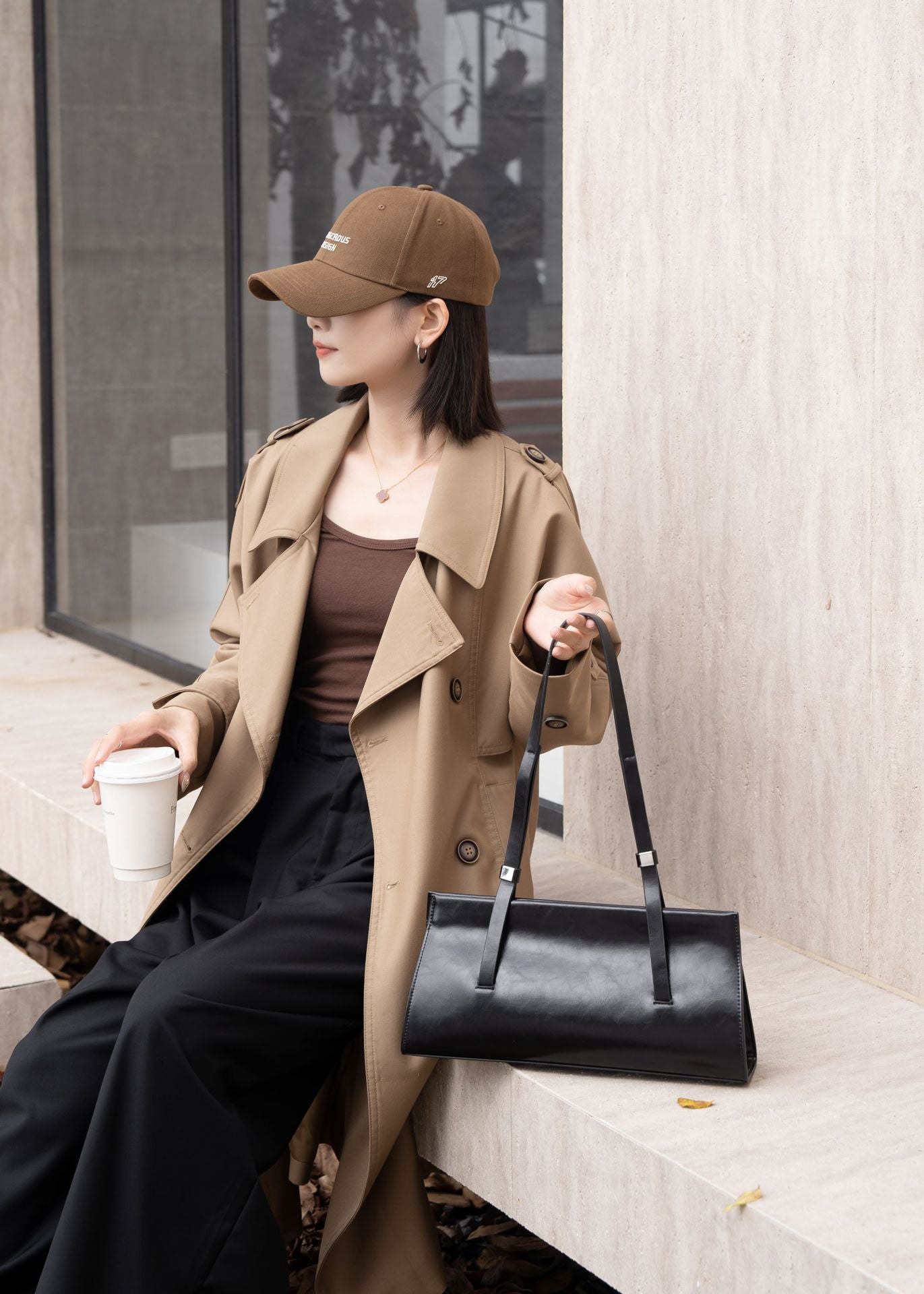 Woman in a trench coat holding a black handbag and a white cup, sitting on a bench.