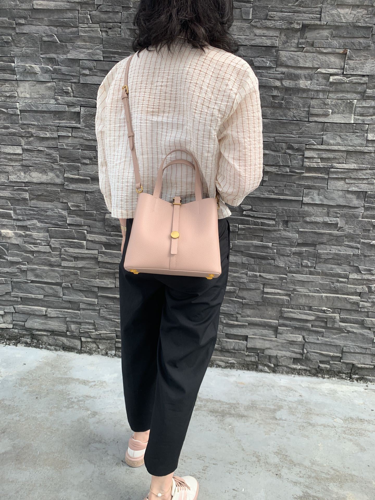 Person wearing a beige top and black pants with a pink handbag against a stone wall.