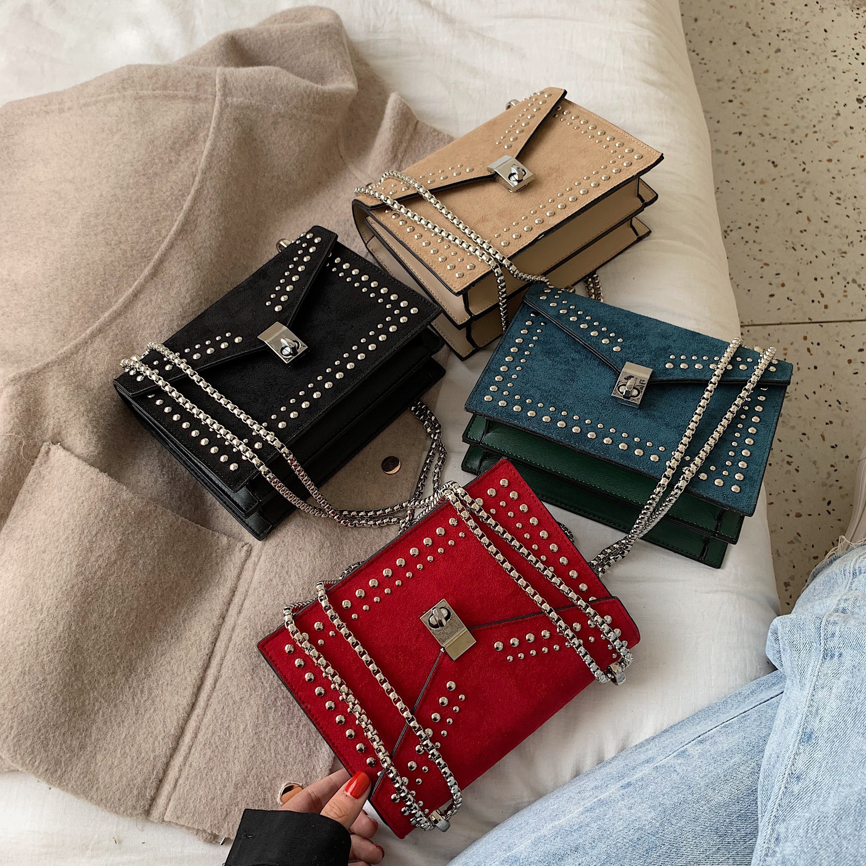 Four small handbags in black, beige, blue, and red with chain details on a beige surface.