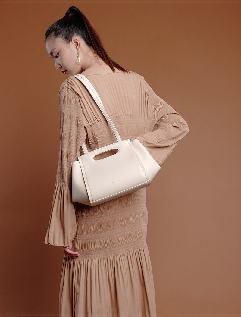 Woman in a beige dress holding a white handbag against a brown background