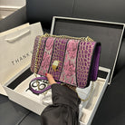 Purple and pink handbag with gold chain held by a hand, displayed in an open box.