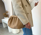 Person holding a woven handbag wearing a beige coat and blue jeans indoors.