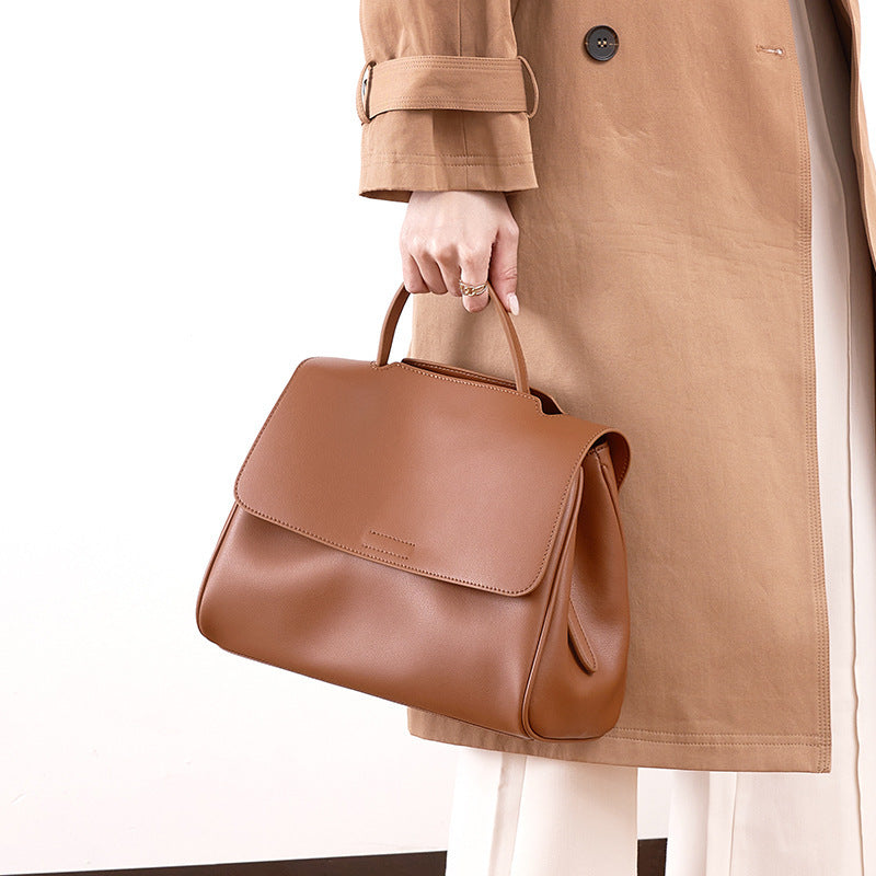 Brown leather handbag held by a person wearing a tan coat on a white background
