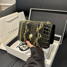 Black handbag with gold accents held over a white box, with a 'THANK' branded bag in the background.