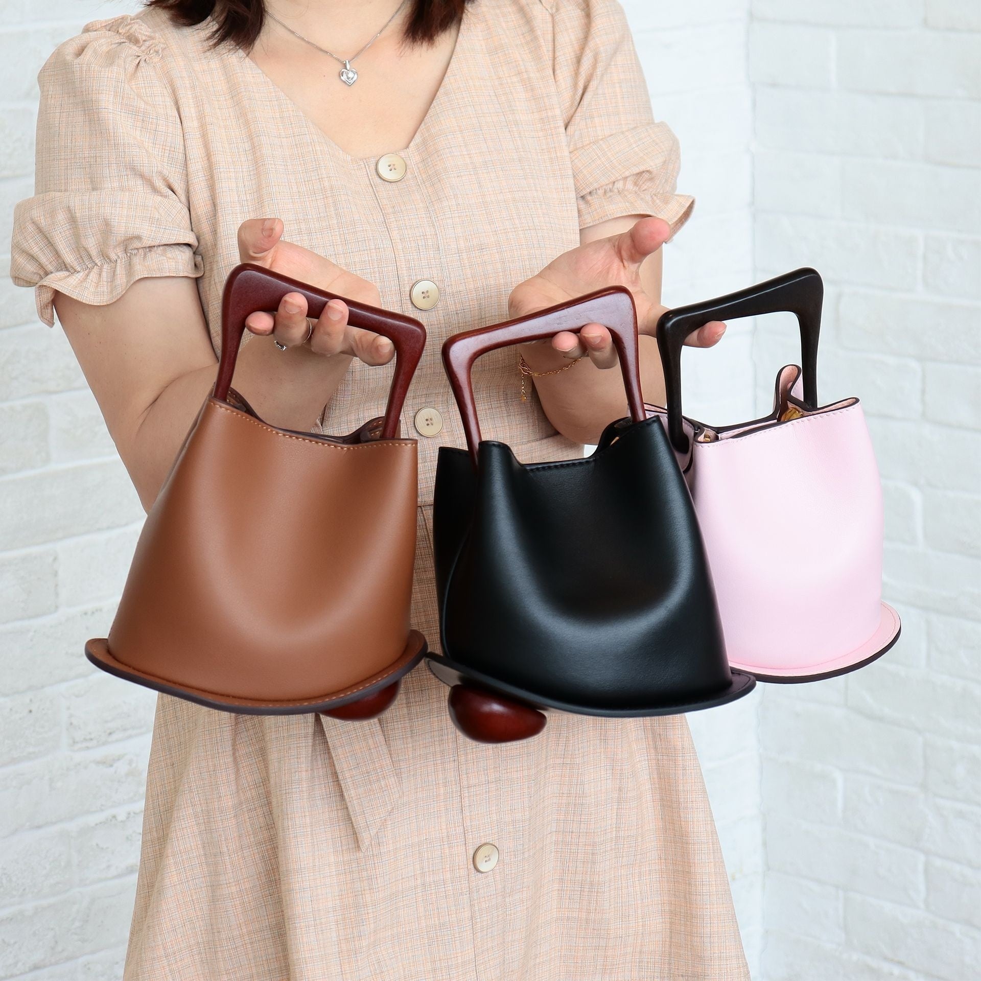 Person holding three different colored handbags against a white brick wall.