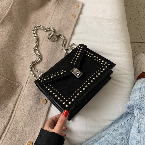 Black studded clutch held by a hand with red nail polish, sitting on a light-colored surface.