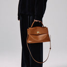 Brown leather handbag held by a person wearing a black outfit on a light gray background