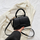 Black handbag held by a hand on a white background with a light-colored fabric.