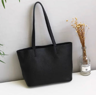 Black handbag on a white surface with green leaves and a vase in the background