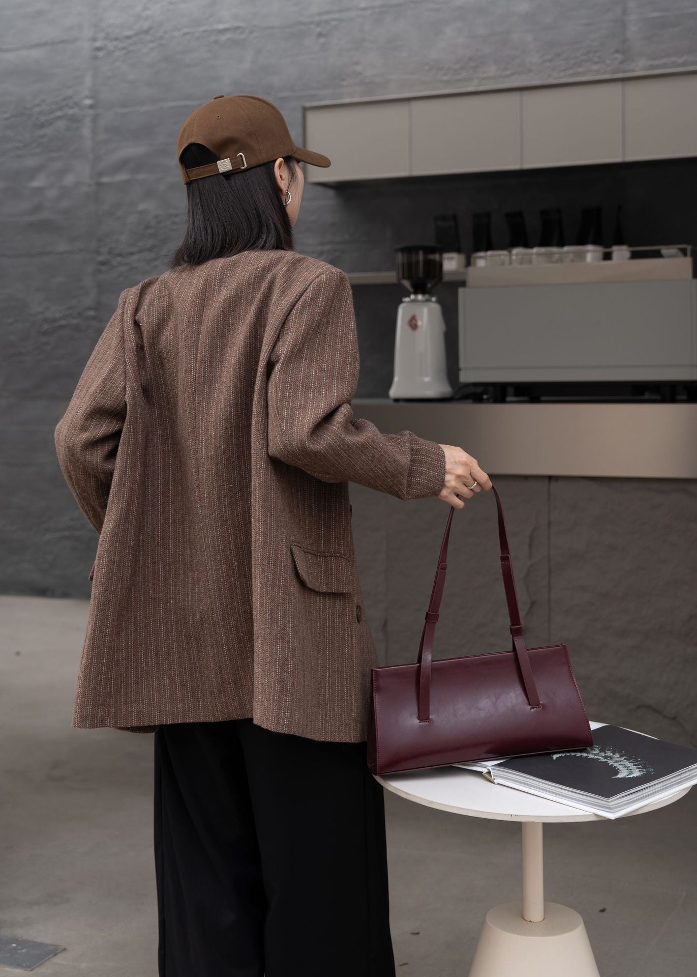 Person in a brown jacket and cap holding a maroon handbag in front of a coffee machine.