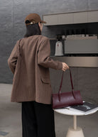 Person in a brown jacket and cap holding a maroon handbag in front of a coffee machine.