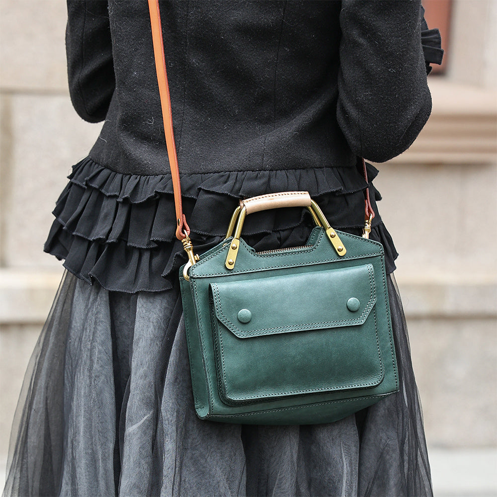 Green handbag with gold accents held by a person wearing a black top and gray skirt.