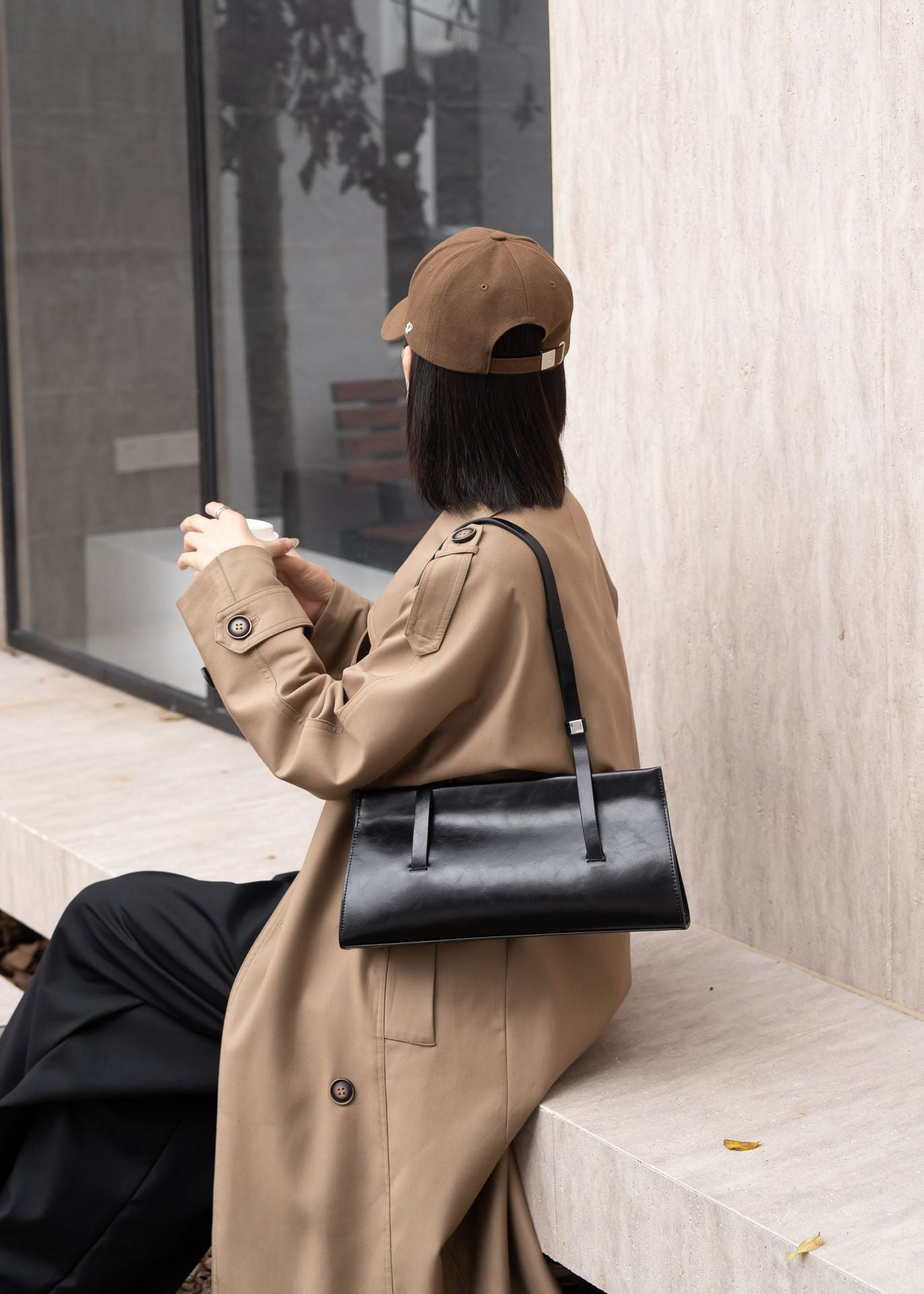 Person wearing a beige coat and cap, holding a black handbag, sitting on a bench.