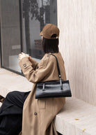Person wearing a beige coat and cap, holding a black handbag, sitting on a bench.