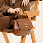 Brown leather handbag held by a person sitting on a wooden chair.
