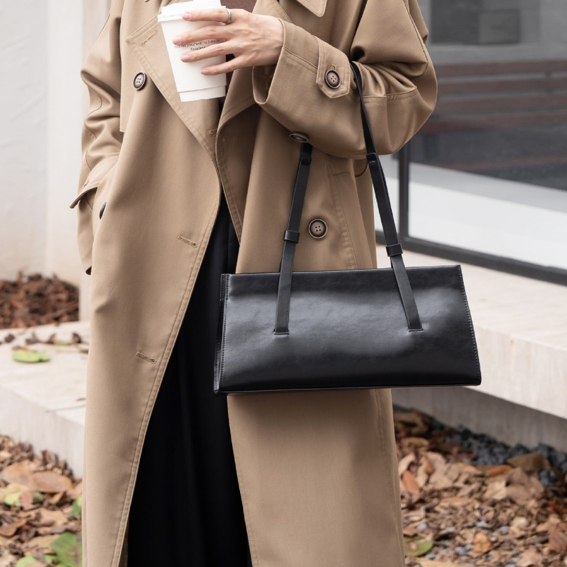 Person wearing a beige trench coat holding a black handbag and a white cup outdoors.