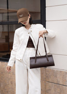 Person wearing a white outfit with a brown cap and holding a brown handbag, standing against a building.