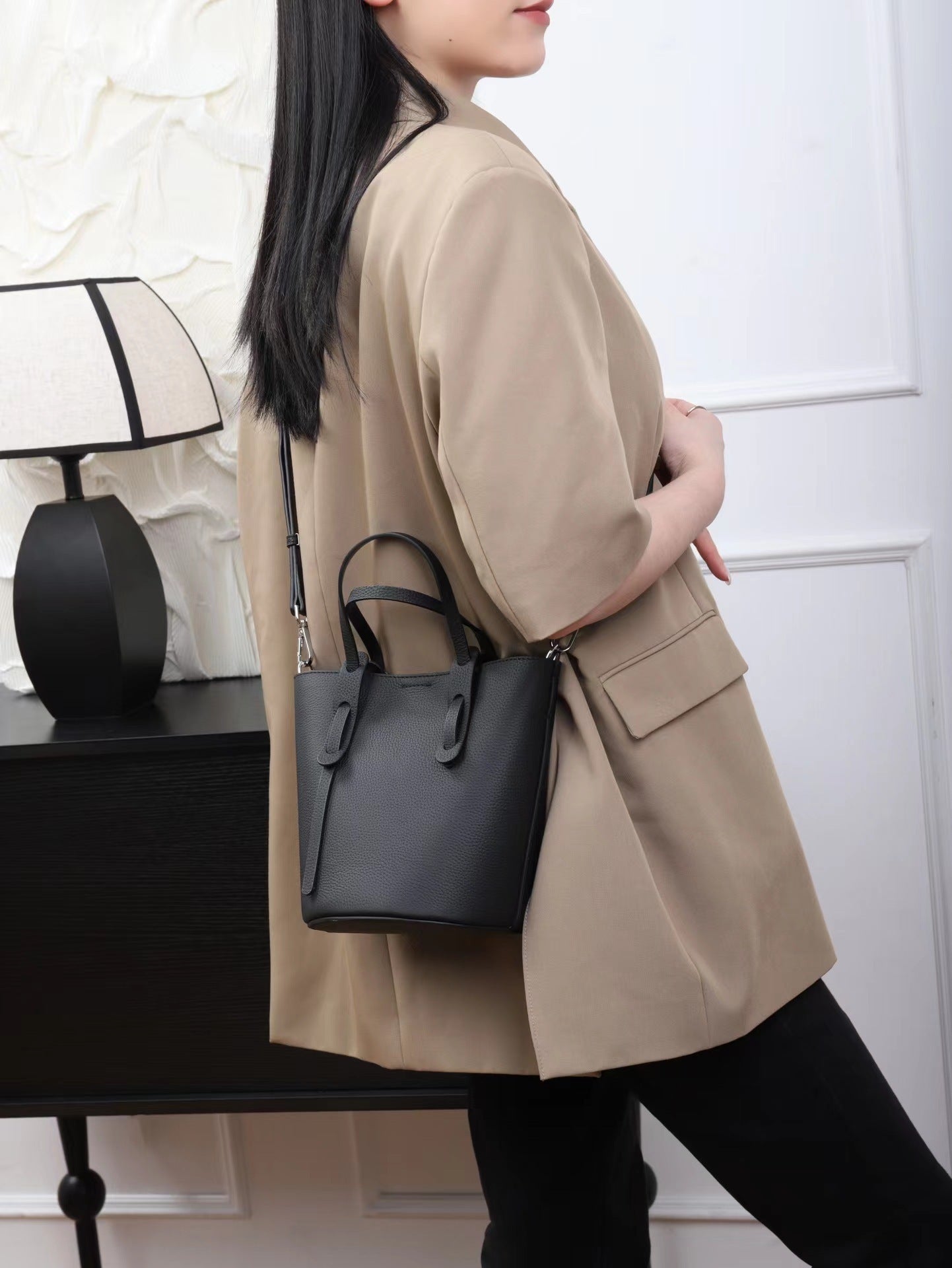 Person wearing a beige coat holding a black handbag in a minimalistic setting.