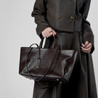 Person holding a dark brown leather handbag against a plain background