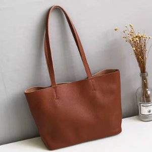 Brown leather tote bag on a white surface with a gray wall and plant in the background