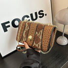 Brown crocodile-patterned handbag with gold chain held by a hand, against a neutral background.