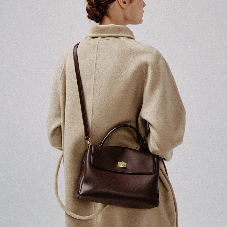 Person wearing a beige coat with a brown leather handbag on a plain background