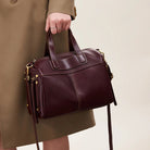 Brown leather handbag held by a person wearing a beige coat on a neutral background
