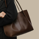 Brown leather tote bag held by a person wearing a black coat on a beige background