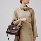 Person wearing a beige coat holding a brown handbag against a plain background