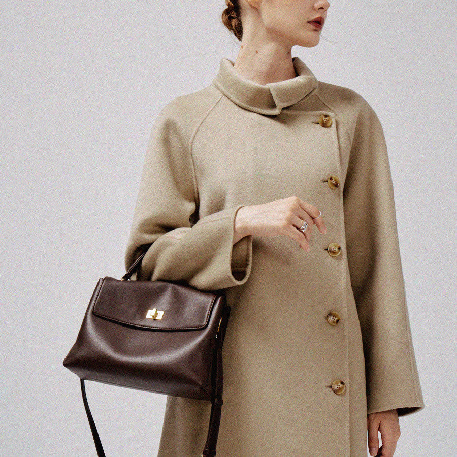 Person wearing a beige coat holding a brown handbag against a plain background