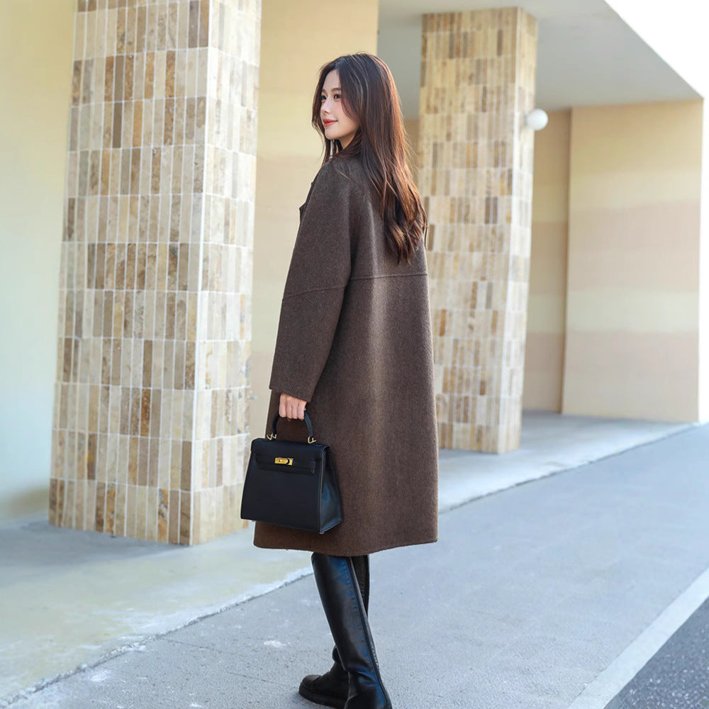 Woman in a brown coat holding a black handbag, standing on a sidewalk.