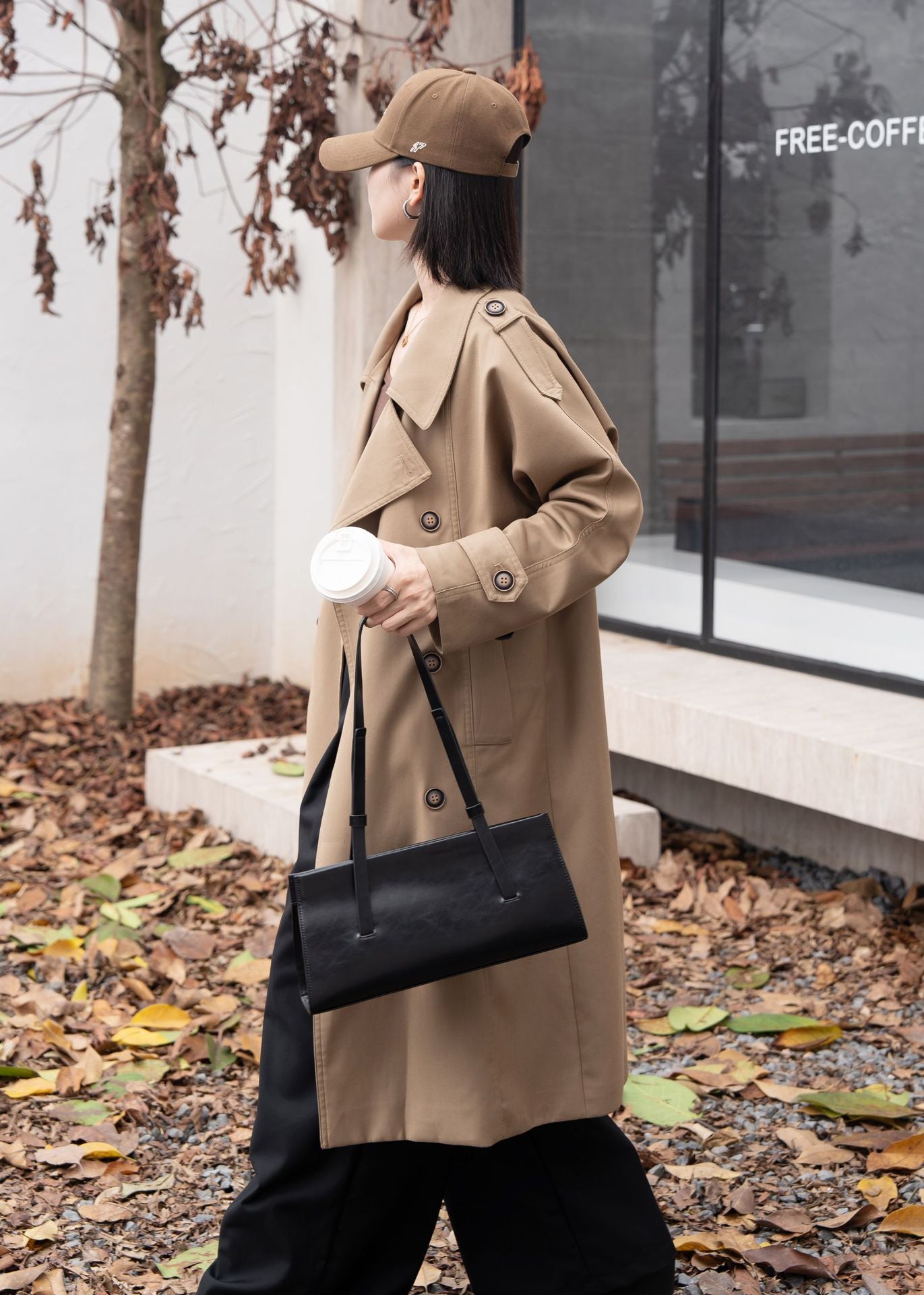 Person wearing a beige trench coat and black cap, holding a black handbag and white cup, standing outdoors.