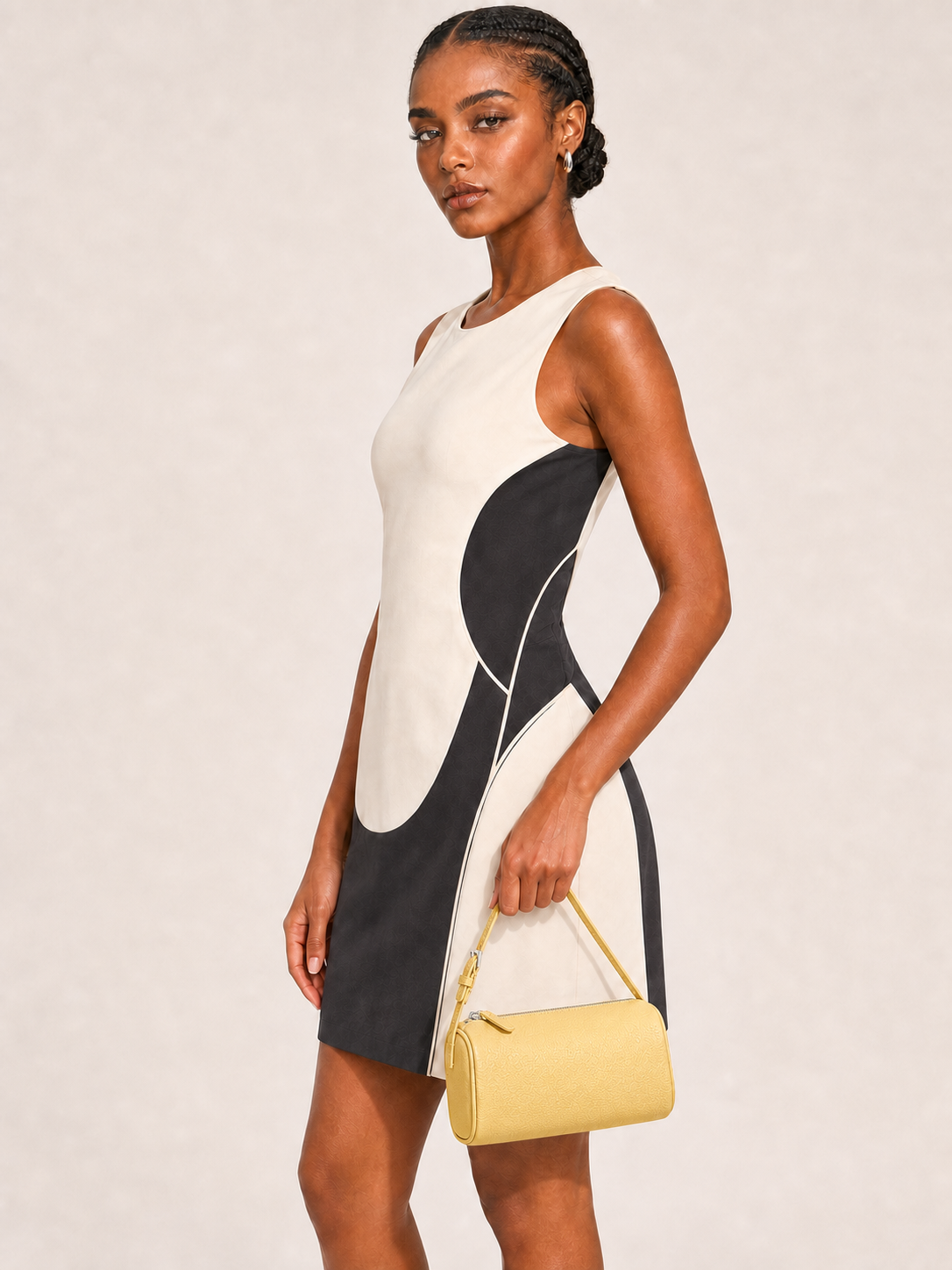 A woman modeling a sleeveless color-block mini dress with a cream, black, and white abstract pattern, holding a small yellow handbag against a plain beige studio background.