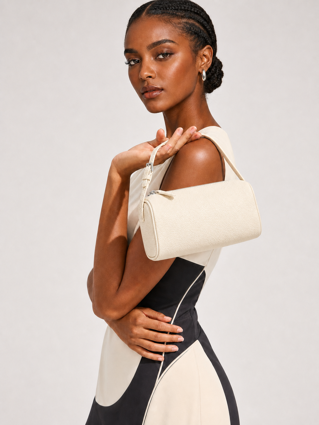 A woman models a cream-colored textured shoulder bag with a top handle, styled with a color-blocked cream and black dress against a light gray studio background.