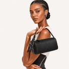 A model with braided hair holds a black textured cylindrical handbag with a black-and-white patterned scarf tied to its handle, against a clean off-white background.