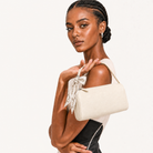 A model with braided hair holds a cream-colored structured shoulder bag with a matching fabric tie detail. She is wearing a black and white top against a clean, off-white studio background.