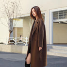 Woman wearing a long brown coat standing outdoors in front of a building.