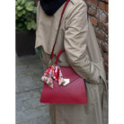 Person holding a red handbag with a floral scarf, wearing a beige coat, against a brick wall.