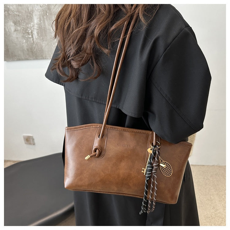 Brown leather handbag with decorative chain strap held by a person wearing a black outfit.