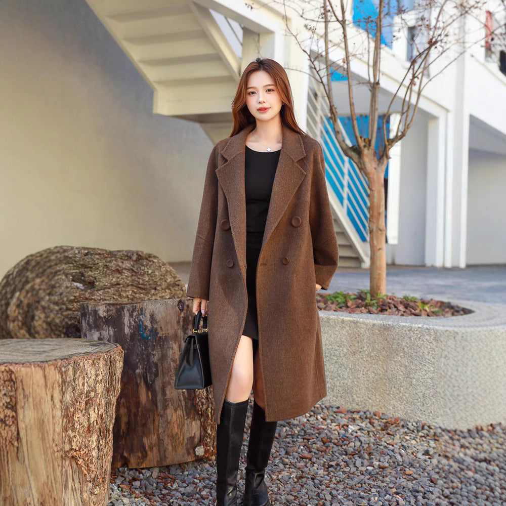 Woman in a brown coat standing outdoors near a tree and concrete structure.