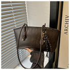 Brown leather handbag with decorative chain held by a hand against a window and book background.