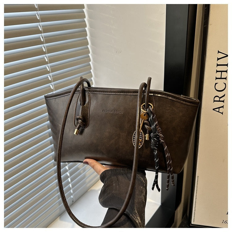 Brown leather handbag with decorative chain held by a hand against a window and book background.