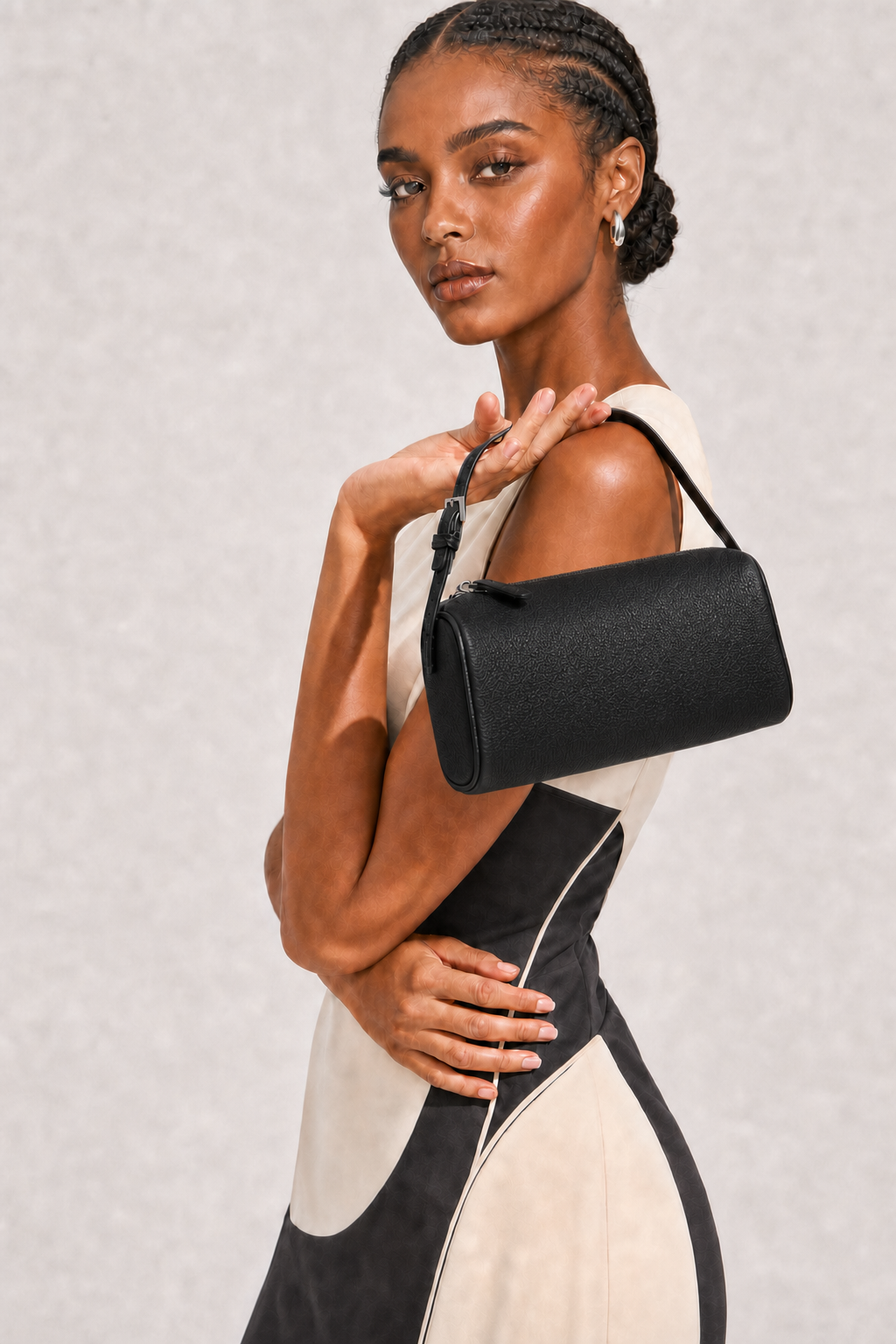 A model wearing a black textured leather cylindrical shoulder bag with a black strap, styled with a black and cream color-blocked dress against a textured light gray background.