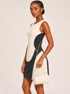 A woman modeling a sleeveless color-block mini dress with a cream and black abstract design, holding a matching cream handbag against a light beige studio background.